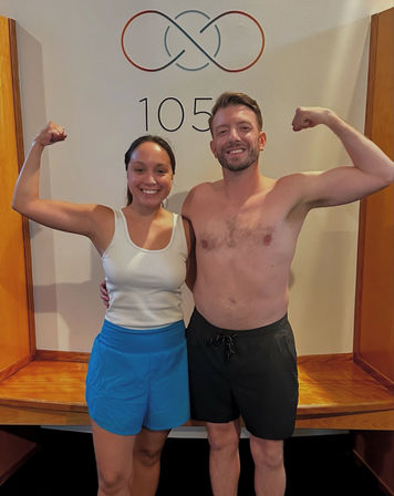Smiling pair flexing biceps after a workout — woman in a white tank and blue shorts and a shirtless man in black shorts posing in front of a wall with a circular logo and the number 105 in a fitness studio.