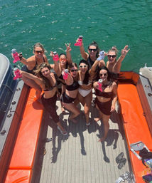 Group of friends in swimsuits cheering with canned drinks on an orange-deck boat over green lake water, sunny summer boat party