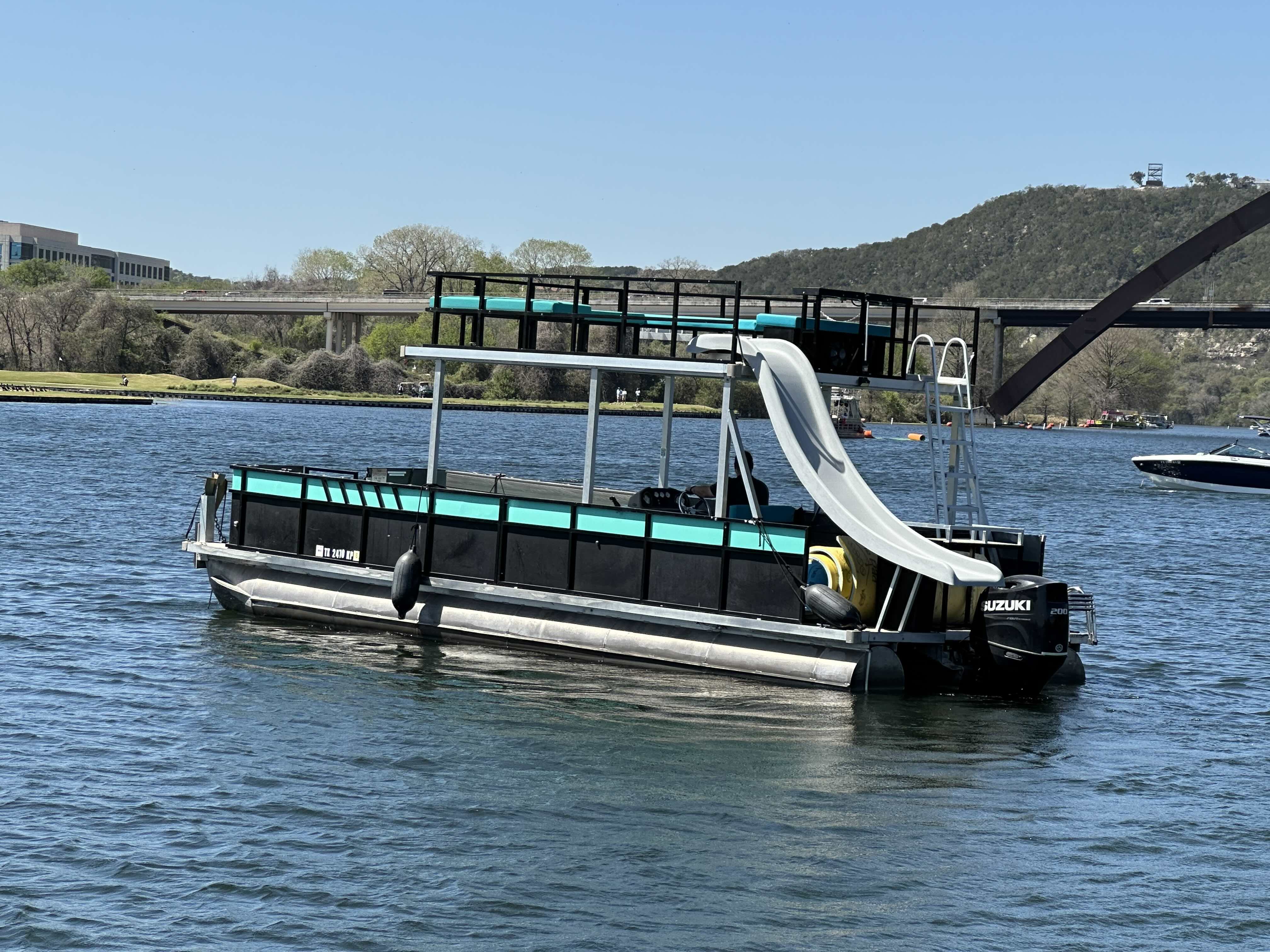 Turquoise-and-black pontoon boat with upper deck and curved white water slide floating on a sunny lake near a bridge and tree-covered hills