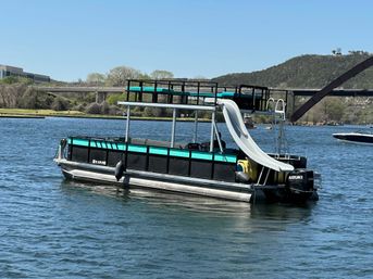 Turquoise-and-black pontoon boat with upper deck and curved white water slide floating on a sunny lake near a bridge and tree-covered hills