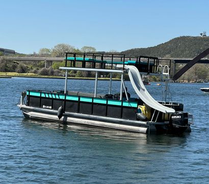 Turquoise-and-black pontoon boat with upper deck and curved white water slide floating on a sunny lake near a bridge and tree-covered hills