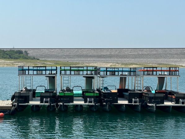 Colorful double-deck pontoon boats moored at a floating dock on a turquoise lake with a distant earthen dam and tree-lined shore.