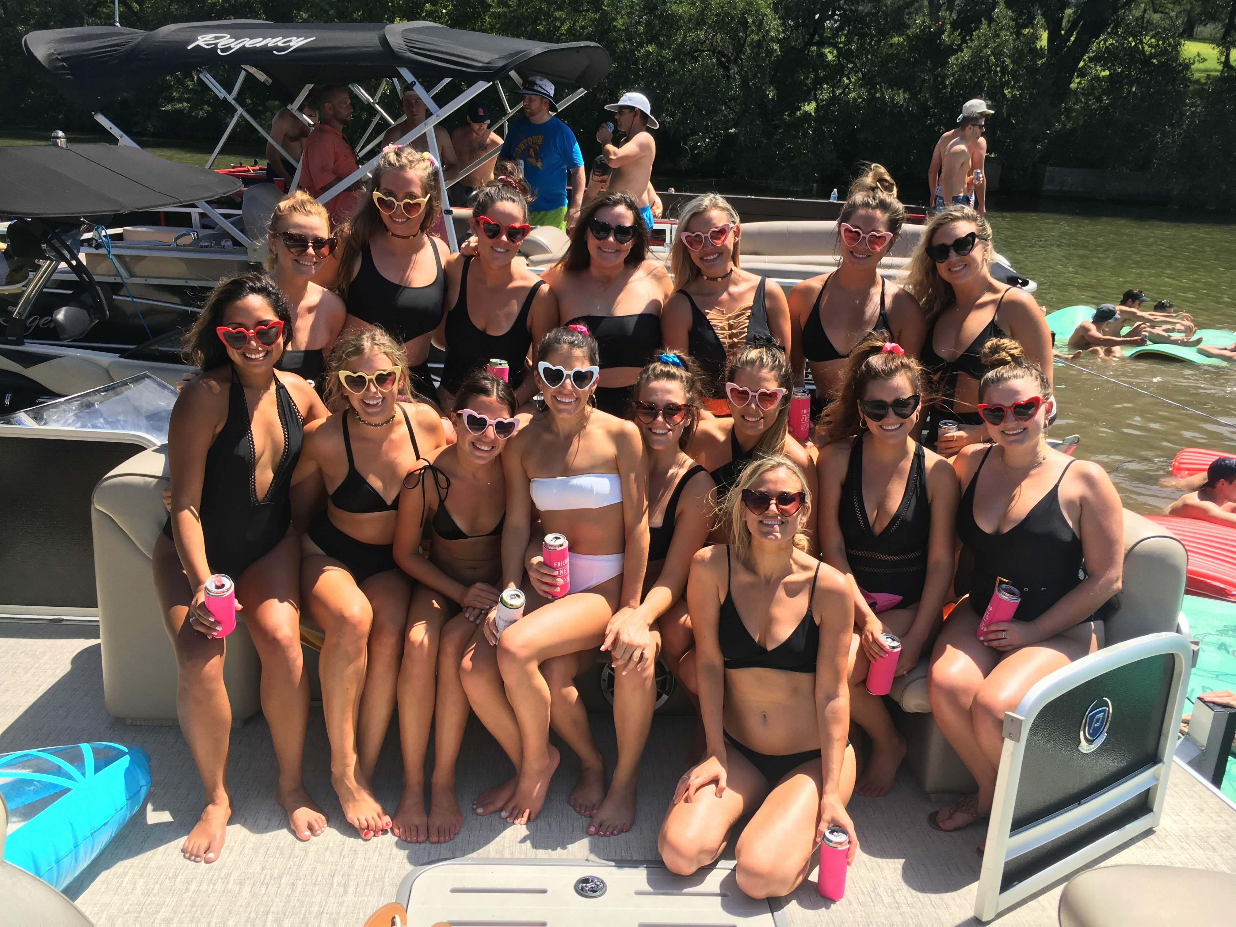 Group of women in swimsuits and heart-shaped sunglasses posing on a pontoon boat at a sunny lake party, holding canned drinks with inflatables and trees in the background.