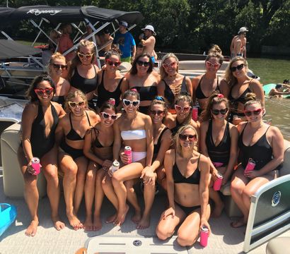 Group of women in swimsuits and heart-shaped sunglasses posing on a pontoon boat at a sunny lake party, holding canned drinks with inflatables and trees in the background.