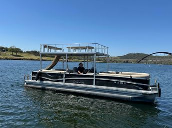 Two-level pontoon boat with rooftop deck and slide cruising on a sunny lake near a distant arched bridge and grassy shoreline under a clear blue sky