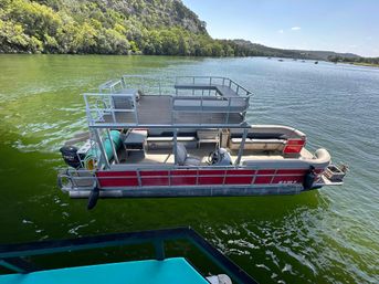 Overhead view of a red double-decker pontoon boat with upper deck, lounge seating and outboard motor floating on a green lake near a wooded shoreline on a sunny day