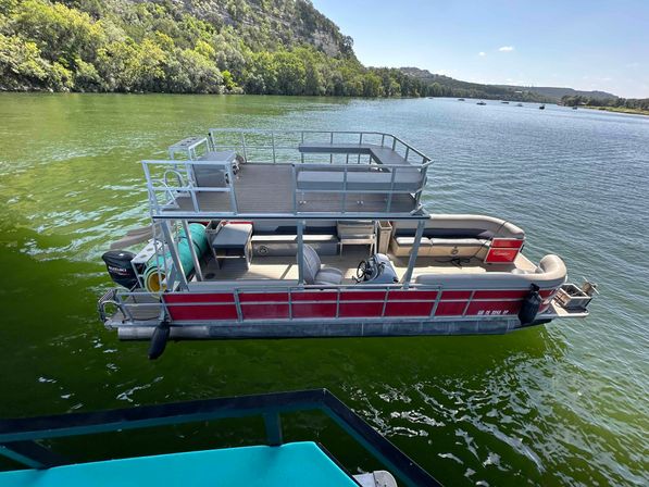 Overhead view of a red double-decker pontoon boat with upper deck, lounge seating and outboard motor floating on a green lake near a wooded shoreline on a sunny day