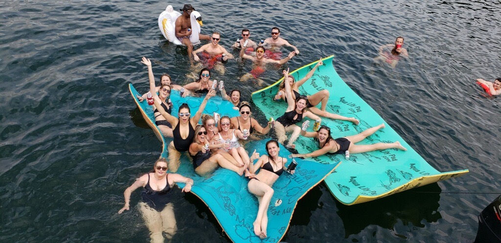 Aerial view of a lively lake party with friends in swimsuits relaxing on turquoise floating mats and an inflatable swan, holding drinks and smiling on sunny summer water.