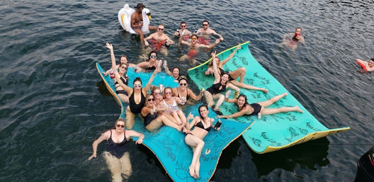 Aerial view of a lively lake party with friends in swimsuits relaxing on turquoise floating mats and an inflatable swan, holding drinks and smiling on sunny summer water.
