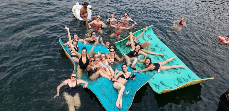 Aerial view of a lively lake party with friends in swimsuits relaxing on turquoise floating mats and an inflatable swan, holding drinks and smiling on sunny summer water.