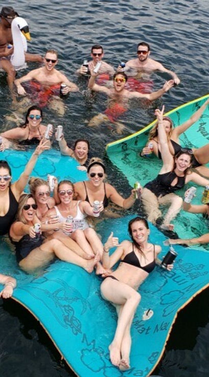 Aerial view of a lively lake party with friends in swimsuits relaxing on turquoise floating mats and an inflatable swan, holding drinks and smiling on sunny summer water.