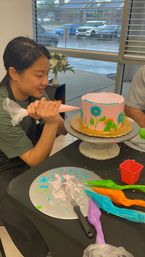 Person piping blue and green floral frosting onto a pink buttercream cake on a rotating stand in a cake-decorating studio, with colorful icing bags, spatula on the table and a rainy street visible through the window.