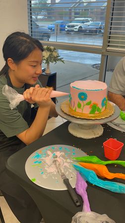 Person piping blue and green floral frosting onto a pink buttercream cake on a rotating stand in a cake-decorating studio, with colorful icing bags, spatula on the table and a rainy street visible through the window.