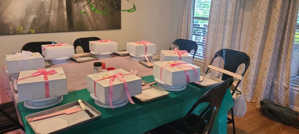 Dining room table set for a cake-decorating party with white cake boxes tied with pink ribbons on turntables, spatulas and trays at each folding chair, light-filled window and curtains