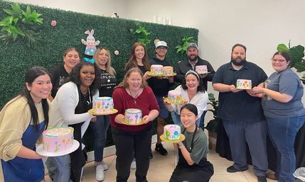 Smiling diverse group holding pastel-decorated cakes at a cheerful cake-decorating class in a studio with a faux green wall and bunny decor.