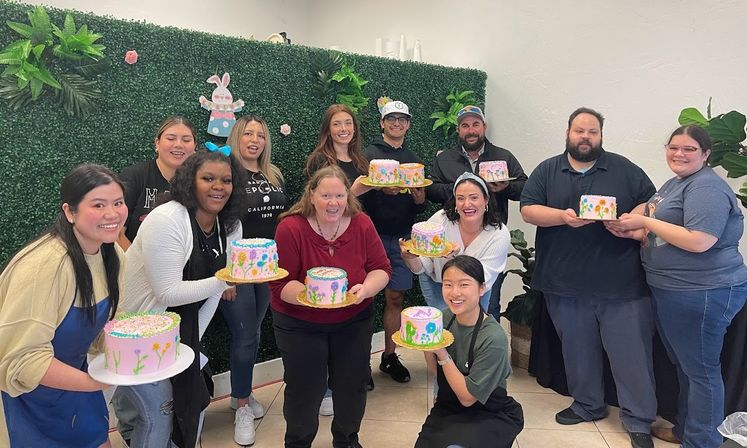 Smiling diverse group holding pastel-decorated cakes at a cheerful cake-decorating class in a studio with a faux green wall and bunny decor.