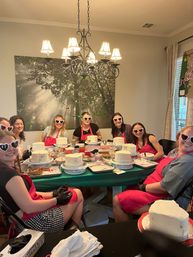 Cake-decorating party in a cozy home dining room: people in red aprons and heart-shaped sunglasses gathered around a table frosting small white cakes under a chandelier