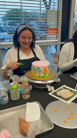 Smiling woman decorating a pink buttercream cake with colorful piped flowers at a cake-decorating workshop, turntable and piping bags on the table.