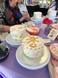 Indoor cake-decorating workshop: hands piping buttercream on mini round cakes with pastel floral designs on turntables, bowls of frosting and a purple tablecloth with a small vase of red roses.