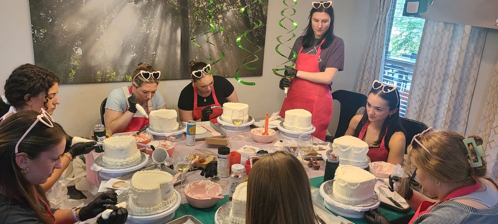 Group of women in aprons and heart-shaped sunglasses decorating white frosted layer cakes around a dining table in a bright home, with bowls of pink frosting, piping bags and sprinkles — cake decorating party/class.