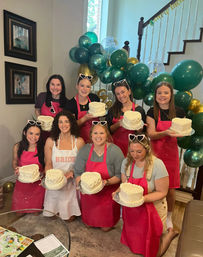 Group of women at a home cake-decorating party wearing pink aprons and heart-shaped sunglasses, each holding a small white frosted cake in front of a green and gold balloon garland by a staircase; one apron reads 'BRIDE'.