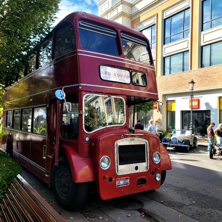 Red vintage double-decker bus parked on a tree-lined city street in a shopping district, with classic cars, benches and storefronts nearby on a sunny day.