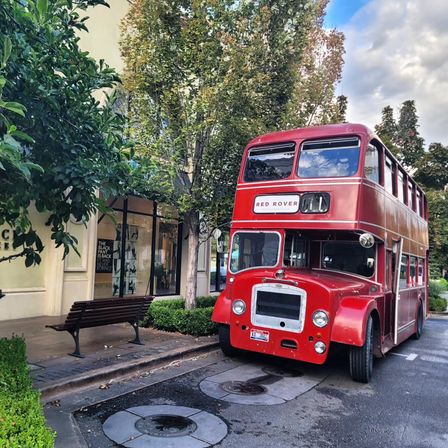 Vintage red double-decker bus labeled "Red Rover" parked on a tree-lined city sidewalk beside a shopfront and wooden bench.