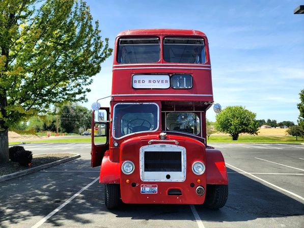 Front view of a vintage red double-decker bus parked in a suburban parking lot next to green trees under a bright blue sky
