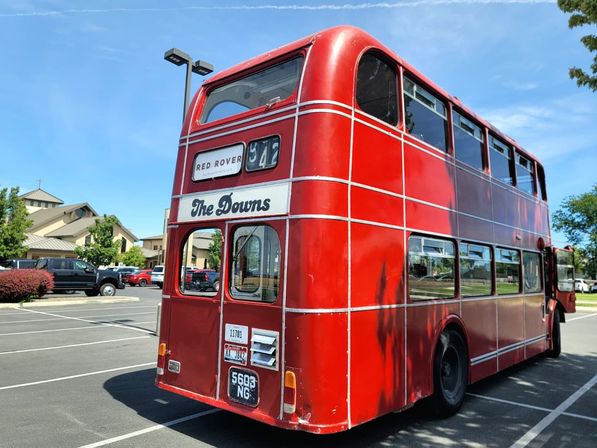 Bright red vintage double-decker bus parked in a suburban parking lot on a sunny day with modern buildings, cars, and trees in the background.