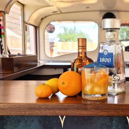 Citrus tequila cocktail on a wooden bar inside a vintage camper — tequila and orange liqueur bottles, whole oranges and a rocks glass with ice and orange slices.
