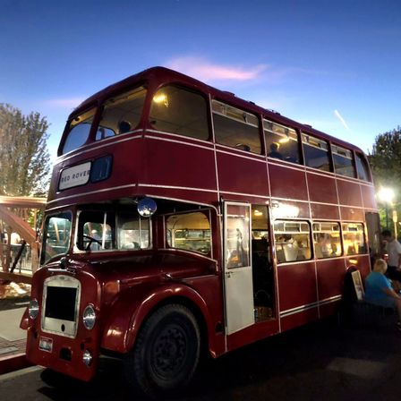 Retro red double-decker bus lit from inside, parked at an evening street festival under a twilight sky with people nearby.
