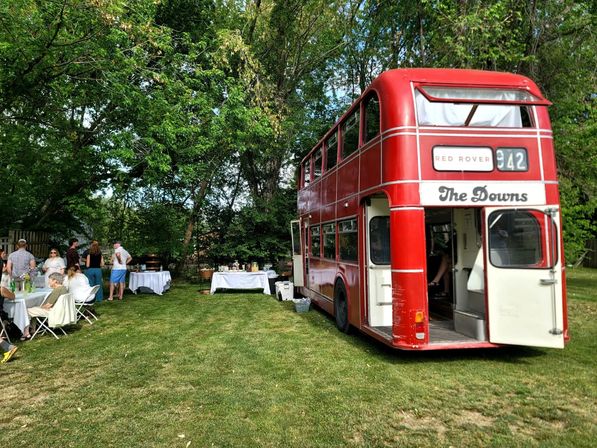 Red vintage double-decker bus parked on a green lawn at a summer outdoor garden party, guests mingling around white-tablecloth tables and a buffet under leafy trees