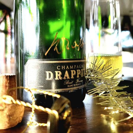 Close-up of a green champagne bottle and a filled flute on a wooden table with a popped cork, twisted wire cage and gold tinsel — a festive celebration scene.