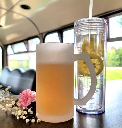 Frosted beer mug with amber drink beside a tall clear tumbler filled with lemon slices and a straw, pink carnation and baby's-breath on a wood table inside a bright vintage trolley.