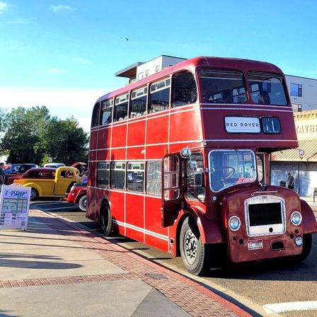 Bright red vintage double-decker bus parked on a sunny downtown street beside colorful classic hot rod cars at an outdoor car show