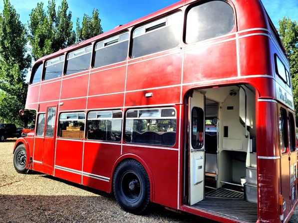 Vintage red double-decker bus parked on gravel under a blue sky, open rear door revealing interior stairs and seating with trees in the background.