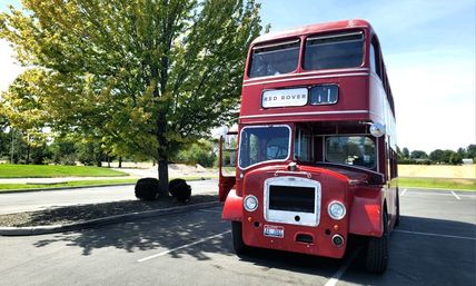 Classic red double-decker bus parked in a sunny suburban parking lot beside a leafy tree and open field under a blue sky.