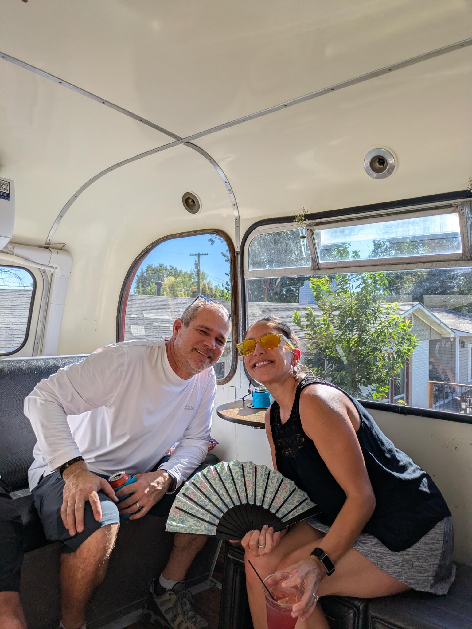 Two adults smiling inside a vintage camper-bus on a sunny day — woman in yellow sunglasses holds a decorative folding fan and pink drink, man in a white shirt holds a canned drink, neighborhood houses visible through the windows.
