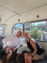 Two adults smiling inside a vintage camper-bus on a sunny day — woman in yellow sunglasses holds a decorative folding fan and pink drink, man in a white shirt holds a canned drink, neighborhood houses visible through the windows.