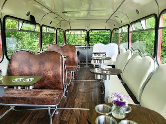Retro bus interior converted into a cozy café with brown leather and white scalloped booths, round wooden tables with stainless cup holders, small purple flower vases, wood-look floor and large windows showing lush green trees.