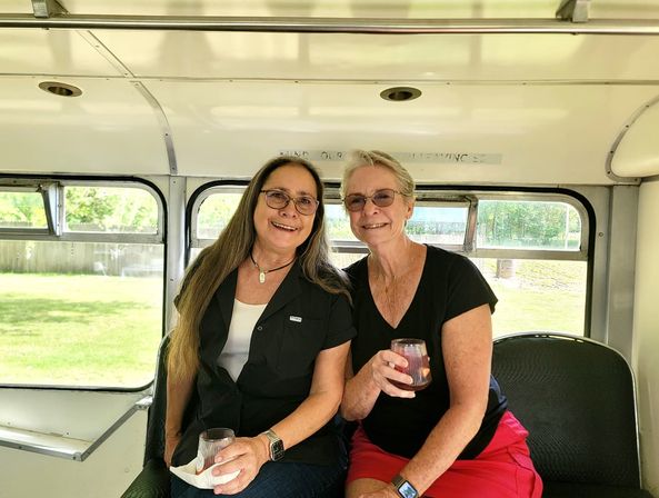 Two smiling women sitting side-by-side inside a vintage bus, each holding a stemless wine glass with sunlight and a green backyard visible through the side windows.