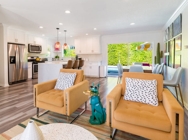 Bright open-plan living room and kitchen in a modern suburban home — mustard-yellow armchairs with spotted pillows, teal geometric dog side table, white kitchen island with copper pendant lights, stainless appliances, dining area and sliding glass doors to a backyard pool and lush hedge.