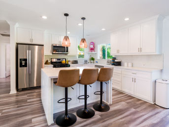 Sunlit modern white kitchen with marble-topped island, three brown swivel bar stools, stainless steel refrigerator, pendant lights, and wood-look vinyl floors