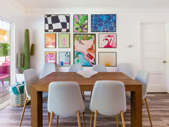Sunny modern dining room with a wooden table and eight gray upholstered chairs, vibrant gallery wall of framed prints, tall cactus by a sliding glass door opening to a bright patio, decorative bowl centerpiece — mid-century modern vibe