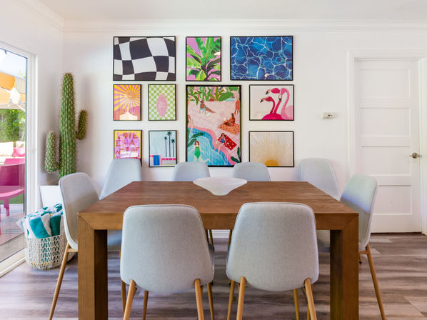 Sunny modern dining room with a wooden table and eight gray upholstered chairs, vibrant gallery wall of framed prints, tall cactus by a sliding glass door opening to a bright patio, decorative bowl centerpiece — mid-century modern vibe