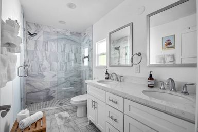 Bright modern marble bathroom featuring a white double-sink vanity with quartz countertop, frameless glass walk-in shower with marble tile walls, chrome fixtures, mirrors and a wooden bench with rolled towels.