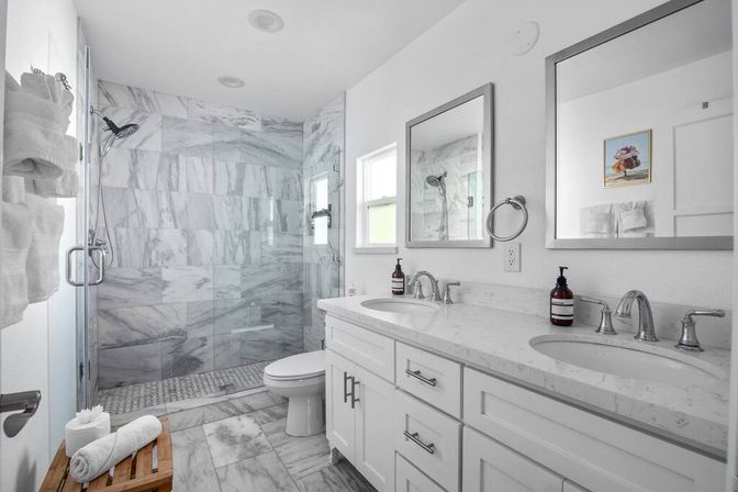 Bright modern marble bathroom featuring a white double-sink vanity with quartz countertop, frameless glass walk-in shower with marble tile walls, chrome fixtures, mirrors and a wooden bench with rolled towels.