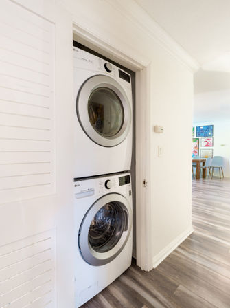 Stacked front-load washer and dryer tucked into a hallway closet with gray wood-look laminate flooring and a bright dining area with colorful wall art visible in the background.