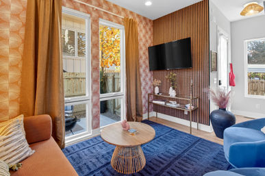Cozy modern living room with wood-slat accent wall and wall-mounted TV, blue textured rug, orange sofa, round rattan coffee table, tall golden curtains and large windows overlooking a fenced backyard with autumn foliage.