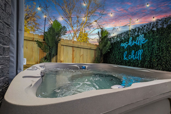 Bubbling outdoor hot tub in a private backyard patio with wooden fence, tropical plants, hanging string lights, blue neon “& chill” sign on a green hedge and a colorful sunset sky